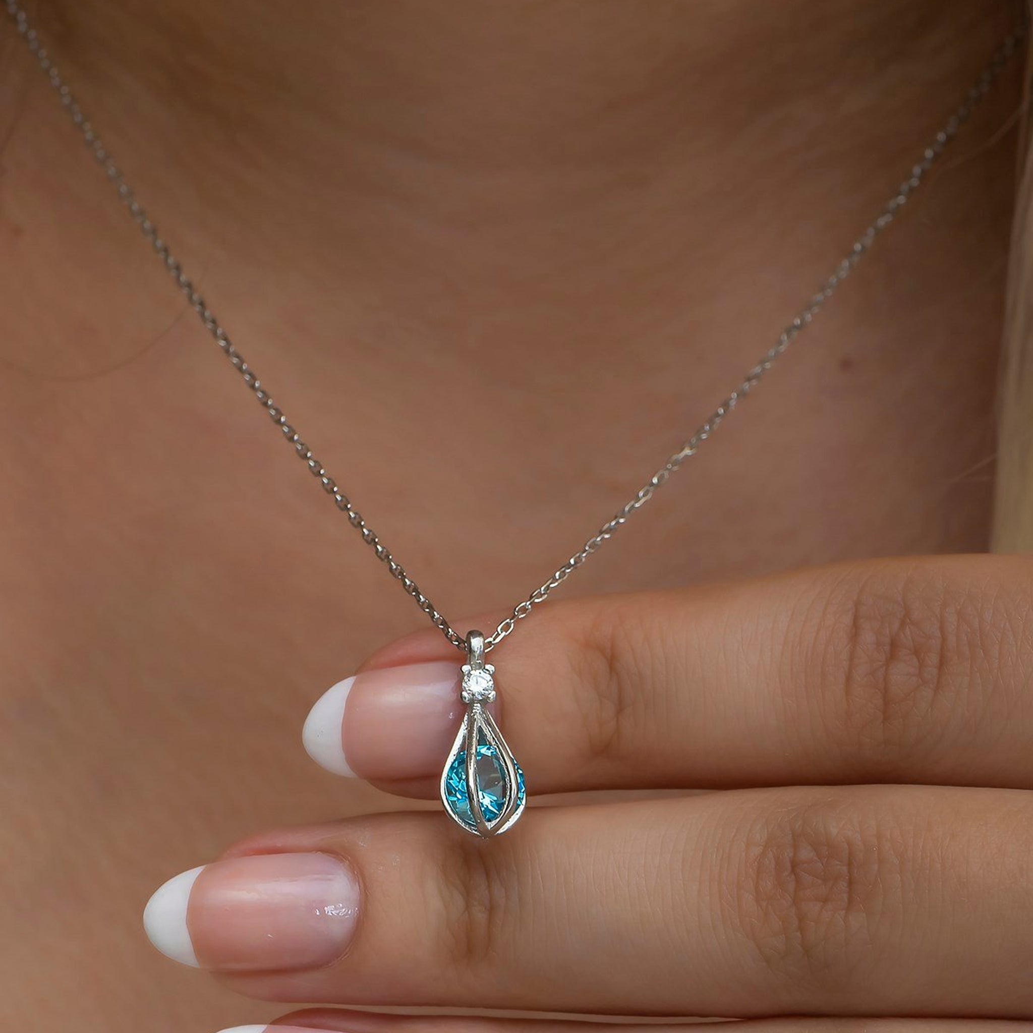 Teardrop-shaped pendant with blue gemstone on a chain held by a hand.