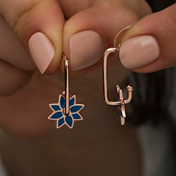 Rose gold earrings with blue floral and cactus designs held by a hand.