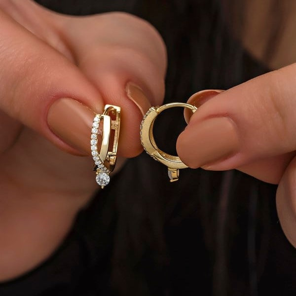 Gold hoop earrings with embedded stones held between fingers against a dark background