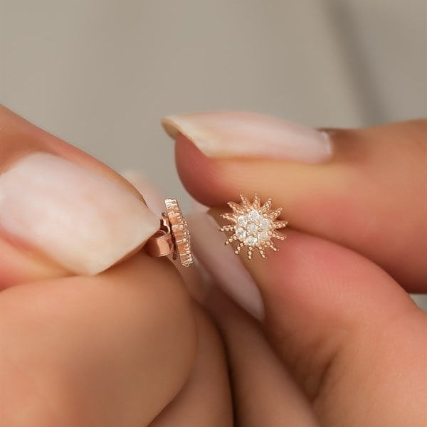Rose gold sun-shaped earrings held between fingers against a neutral background