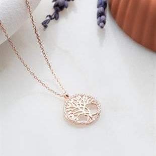 Rose gold tree of life necklace on a white surface with purple flowers in the background