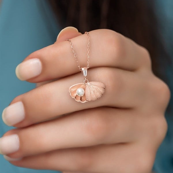 Rose gold necklace with a shell and pearl pendant held by a hand against a blurred background