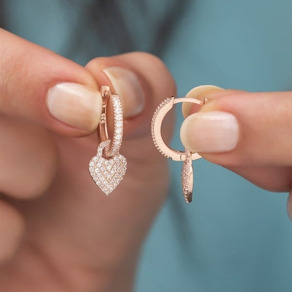 Rose gold hoop earrings with heart-shaped pendants held by a person against a blurred background