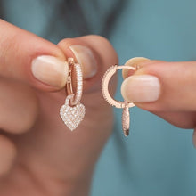 Rose gold hoop earrings with heart-shaped pendants held by a person against a blurred background