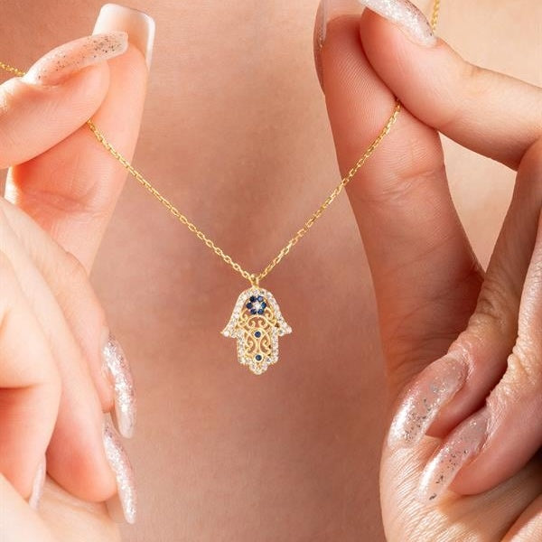 Gold necklace with a hamsa hand pendant held by hands with glittery nail polish.