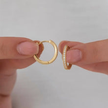Two gold hoop earrings held between fingers against a neutral background