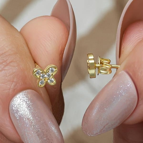 Gold butterfly earrings held between fingers with a neutral background