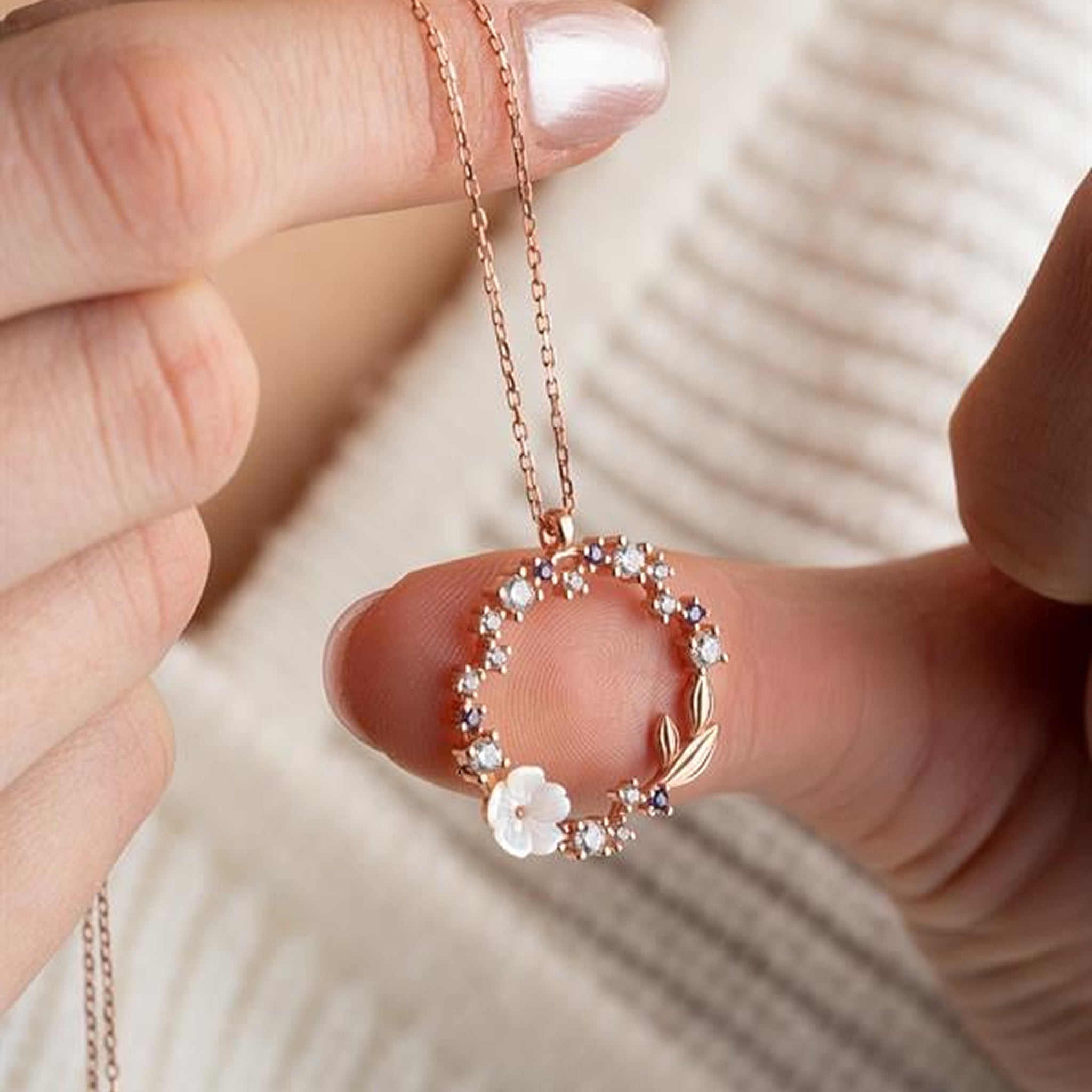 Rose gold necklace with floral pendant held by a hand against a neutral background