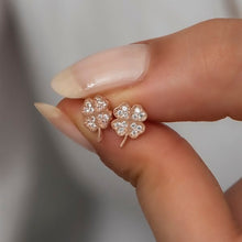 Rose gold four-leaf clover earrings held between fingers against a neutral background