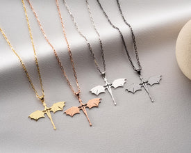 Four bat-shaped pendants on chains in gold, rose gold, silver, and black on a light gray background.