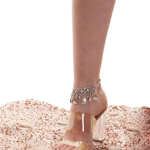 Clear high-heeled sandal with decorative anklet on a sandy background