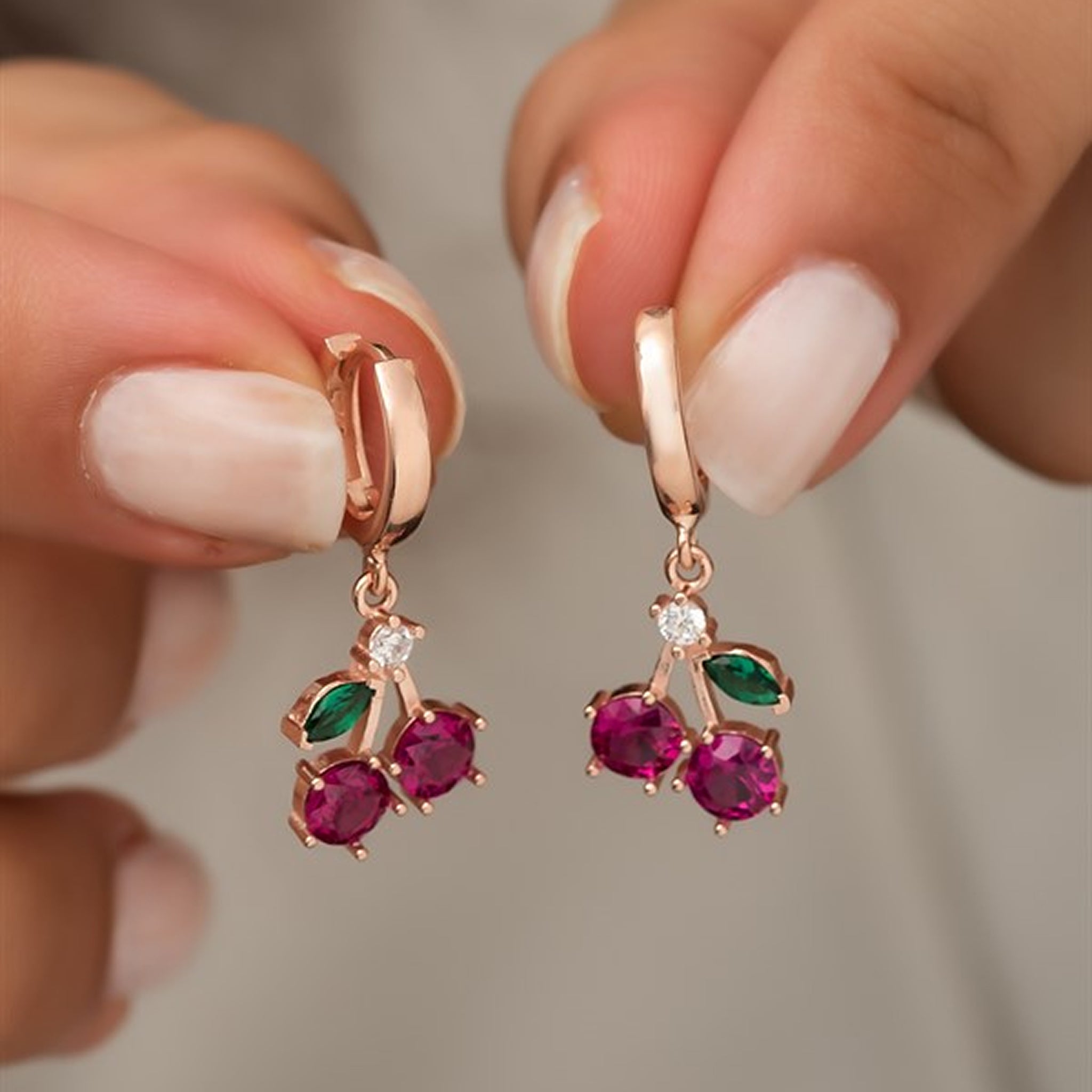 Rose gold earrings with red gemstones held by a hand against a neutral background