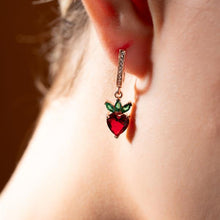 Close-up of a heart-shaped earring with red and green stones worn by a person.