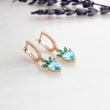 Rose gold earrings with heart-shaped blue and green gemstones on a light background