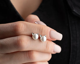 Cat-shaped pearl earrings held between fingers against a dark background