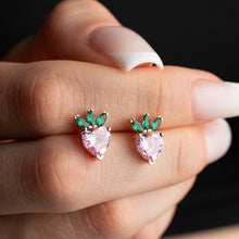 Heart-shaped pink gemstone earrings with green accents held between fingers against a dark background