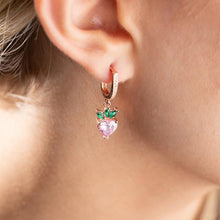 Close-up of a pink heart-shaped earring with green leaves worn by a person.
