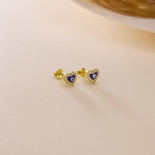 Gold heart-shaped earrings with blue evil eye design on a beige background