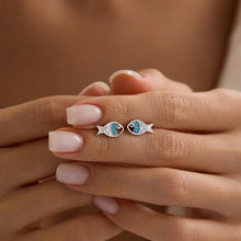 Fish-shaped earrings with blue gemstones held between fingers against a neutral background