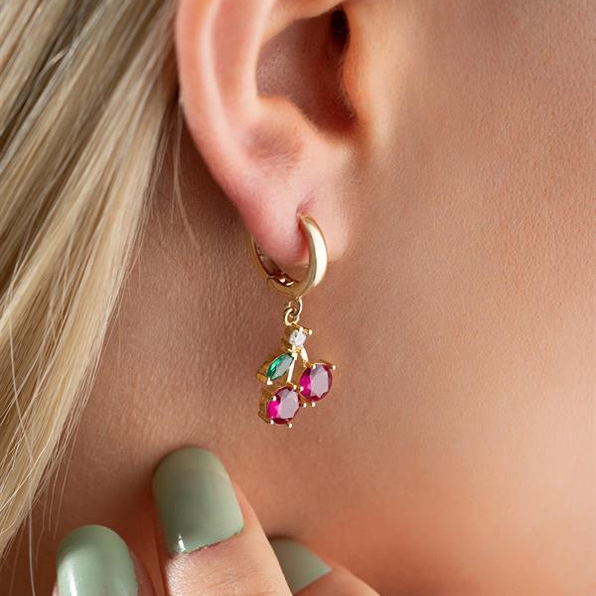 Gold earring with colorful gemstones worn by a person with light skin and green nail polish.