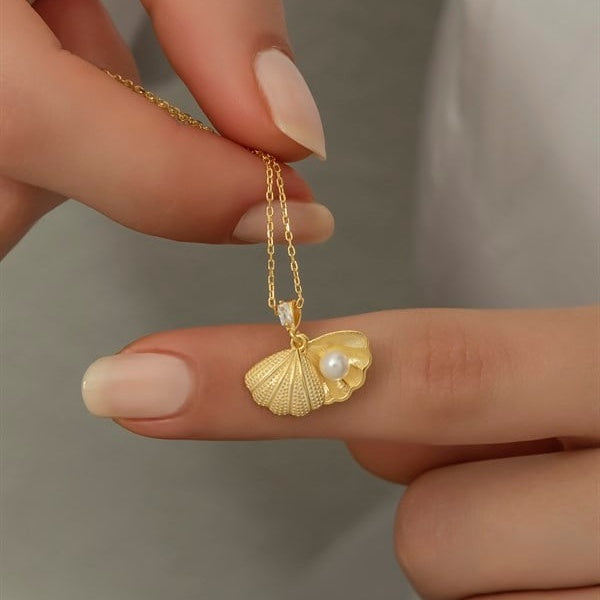 Gold necklace with a shell and pearl pendant held by a hand against a neutral background
