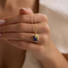 Hand holding a gold necklace with a blue pendant against a neutral background