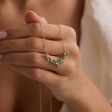 Gold necklace with green gemstones held in a hand against a neutral background