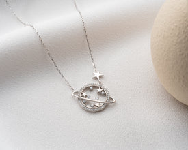 A celestial jewelry flat-lay featuring the sterling silver Saturn necklace alongside moon and star pendants.