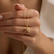 Hand holding a gold necklace with a pink pendant against a neutral background