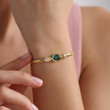 Gold bracelet with a green gemstone on a wrist against a soft pink background