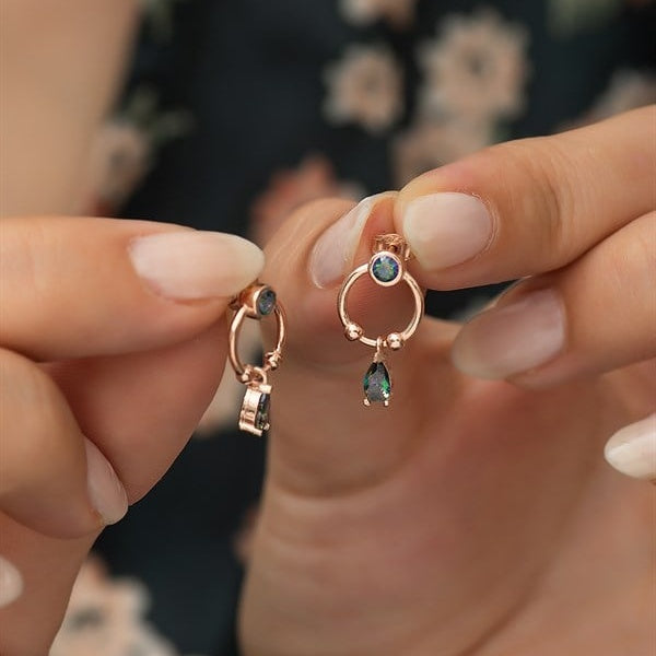 Rose gold earrings with green gemstones held between fingers against a blurred background