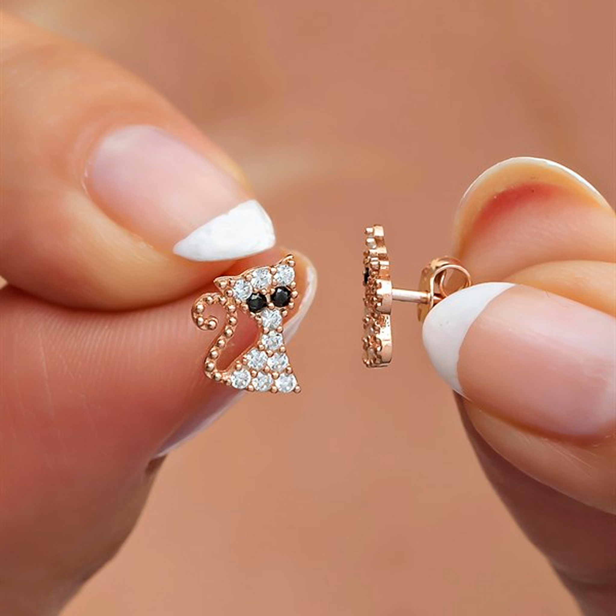 Rose gold cat-shaped earrings held between fingers with a blurred background