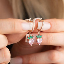 Hand holding a pair of strawberry-shaped earrings with pink gemstones and green leaves.