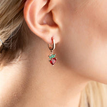 Close-up of an ear wearing a heart-shaped earring with red and green stones.