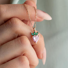 Heart-shaped pink gemstone pendant with green accents on a rose gold chain held by a hand.