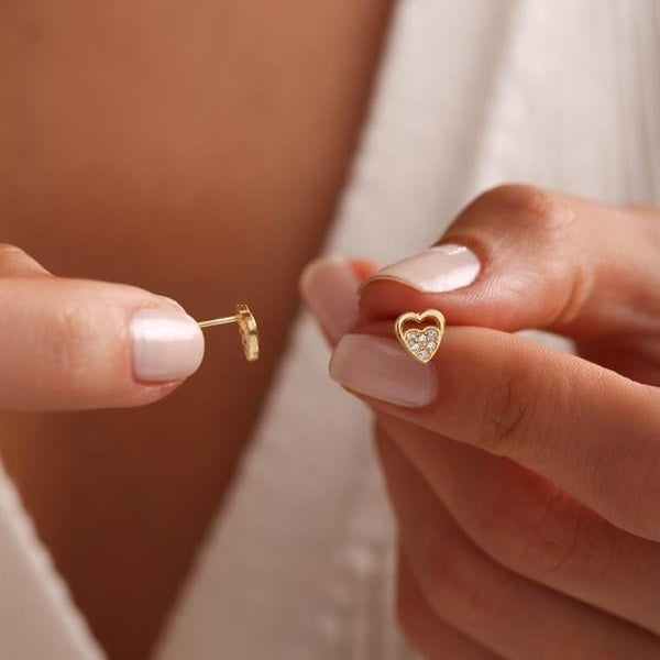 Gold heart-shaped earring held between fingers against a neutral background