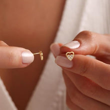 Gold heart-shaped earring held between fingers against a neutral background
