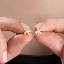 Gold star-shaped earrings held between fingers against a neutral background