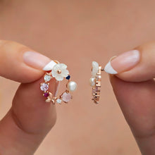 Rose gold earrings with floral and gemstone designs held by a hand against a beige background