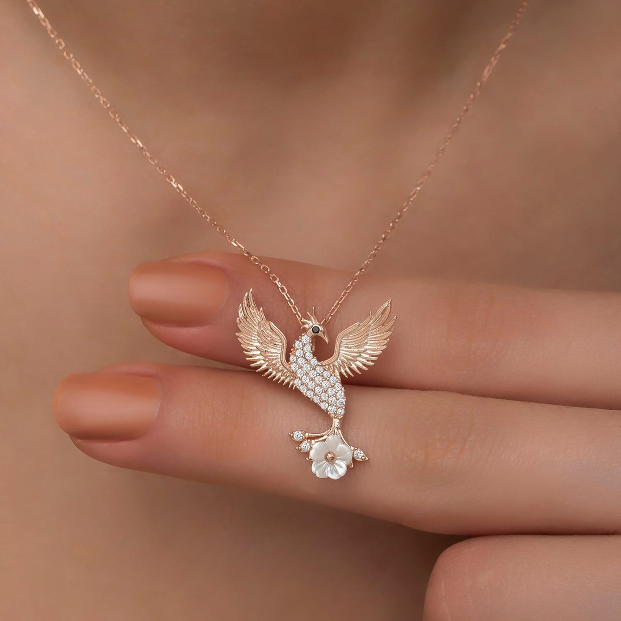 Rose gold necklace with a phoenix pendant held by a hand against a neutral background