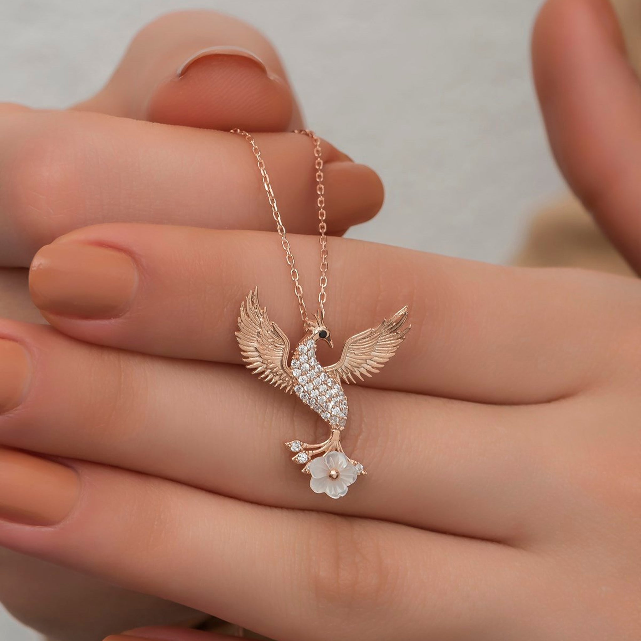Rose gold phoenix necklace with diamond details held in a hand against a neutral background