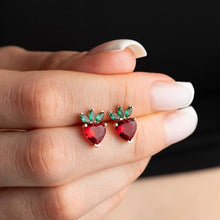 Heart-shaped earrings with red stones and green accents held between fingers against a dark background