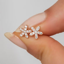 Rose gold flower-shaped earrings with clear stones held between fingers against a neutral background
