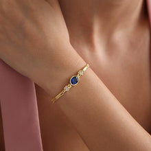 Gold bracelet with a blue gemstone on a wrist against a pink fabric background