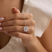 Hand holding a silver necklace with a flower pendant against a neutral background