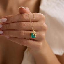 Hand holding a gold necklace with a green pendant against a neutral background