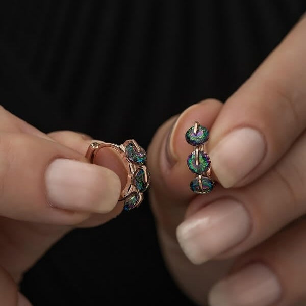 Close-up of hands holding a pair of rose gold hoop earrings with multicolored gemstones against a dark background.